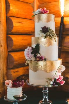 three tiered wedding cake with flowers on each layer and gold leaf decoration, in front of rustic log wall