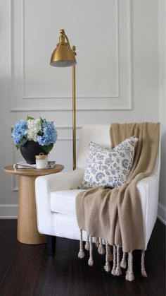 a white chair with a blanket on it next to a lamp and flowers in a vase