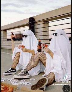 two women dressed in white are sitting on the ground with their legs crossed and holding beer