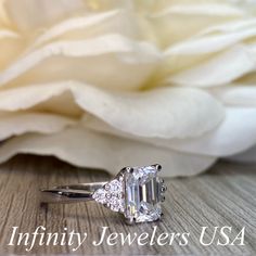 an engagement ring with a large diamond surrounded by smaller diamonds on a table next to a white flower