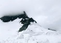 a mountain covered in snow and clouds on top of it's side, with a single person standing at the bottom