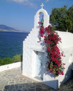 a white church with red flowers growing on it's side and the ocean in the background