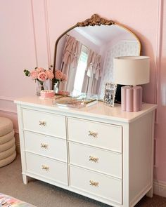 a white dresser topped with a mirror next to a lamp and a pink flower vase