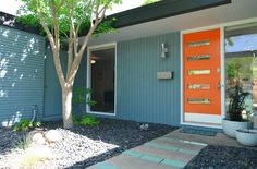 an orange front door on a blue house with gravel and trees in the foreground