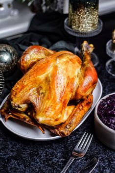 a roasted turkey sits on a plate next to purple cabbage and silverware, along with other holiday foods