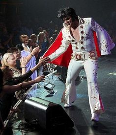 a man in white elvis suit standing on stage with his hands out to the crowd