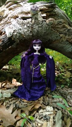 a doll is sitting under a tree in the woods, wearing a purple outfit and long hair