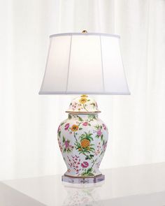 a lamp that is sitting on top of a white table with a flowered vase underneath it