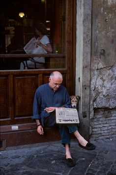 a man sitting on the sidewalk reading a newspaper with his dog in front of him