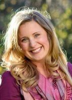 a woman with long blonde hair wearing a purple shirt and smiling at the camera while standing in front of some trees