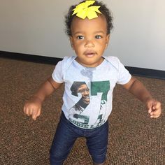 a baby girl with a yellow bow on her head standing in front of a wall