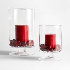 two clear glass vases with red candles and berries in them on a white surface