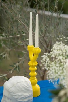 two yellow candles sitting on top of a blue table next to white flowers and greenery