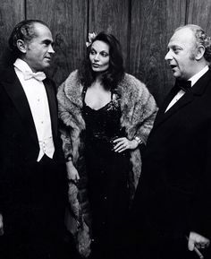 two men and a woman standing next to each other in formal wear, one wearing a fur coat