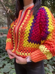 a woman wearing a multicolored crocheted jacket standing in front of trees