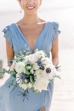 a woman in a blue dress holding a bouquet of white flowers and greenery on the beach