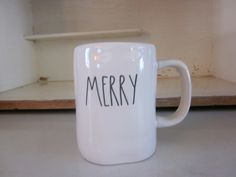 a white coffee mug with the word merry written in black ink on it, sitting next to a shelf