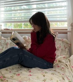 a woman sitting on a bed reading a book in front of a window with blinds