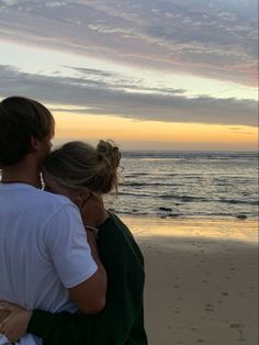 a man and woman are hugging on the beach at sunset, with water in the background