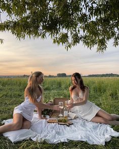 two women in white dresses are sitting on a blanket and having a picnic at the park