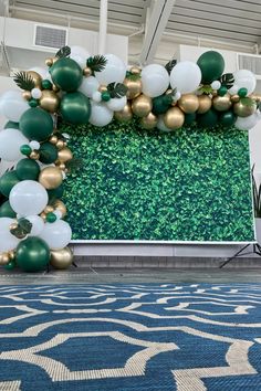 a large green and gold balloon arch in an indoor space with blue carpeting, white balloons and greenery on the wall