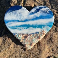 a heart shaped glass piece sitting in the sand on top of a sandy beach with blue sky and clouds painted on it