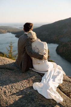 a bride and groom sitting on top of a mountain looking out at the valley below