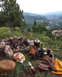 there are many items on the ground near an open fire in the mountains, including bread and jams