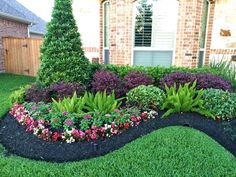 a flower bed in front of a brick house with green grass and flowers around it