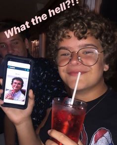 a young man holding up a cell phone with a drink in front of him