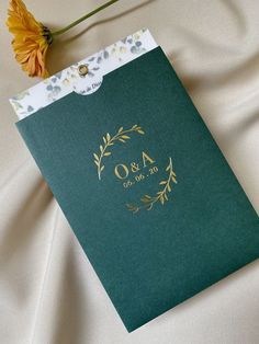 a green and gold wedding guest book on a white bed sheet next to a yellow flower