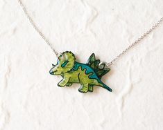 This fantastic jurassic triceratops pendant is hand-illustrated in shades of green and teal with a glossy finish. The pendant hangs on a fine silver gold chain. It measures about 3 cm wide and is coated in resin. The chain is 16K gold plated over brass and adjusts from 16-18 inches in length.Find matching earrings here: https://www.etsy.com/listing/741303409/triceratops-dinosaur-stud-earrings-dino+++ Caring for your necklace +++- Avoid contact with perfumes, body oils, and other chemicals, inclu Dino Necklace, Dinosaur Jewelry, Dinosaur Pendant, Body Oils, Special Jewelry, Barbie Friends, Household Cleaners, Jewelry Cleaner, Hand Illustration