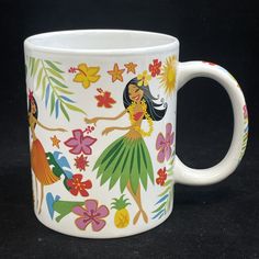 a white coffee mug decorated with hawaiian hula girls and pineapples, on a black background