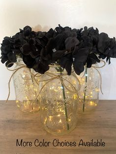 three mason jars filled with black flowers and fairy lights are sitting on a wooden table