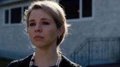 a woman with blonde hair wearing a black shirt and pearl necklace in front of a house