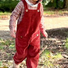 a young child wearing red overalls and a hat walking in the grass with a stick