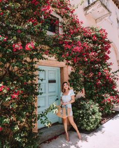 a woman standing in front of a blue door with pink flowers on the outside and green doors