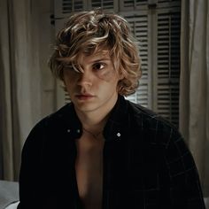 a young man with curly hair sitting in front of a window looking at the camera