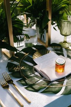 a table set with gold place settings, greenery and a jar of honey on it