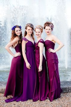 three women in purple dresses standing next to a fountain with water cascading behind them