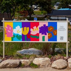 a sign with flowers painted on it in front of some rocks and grass near trees