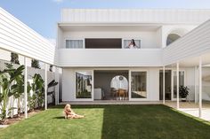 a woman sitting on the grass in front of a house with white walls and windows