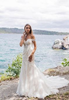 a woman in a wedding dress standing by the water with her hand on her ear