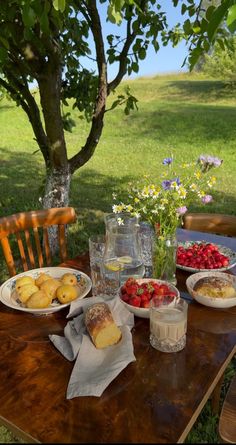 a wooden table topped with plates of food next to a forest filled with flowers and trees