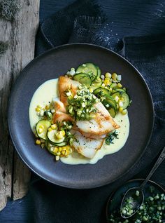 a black plate topped with fish covered in sauce next to cucumbers and corn