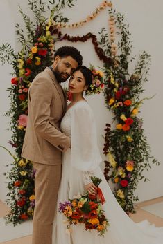 a bride and groom standing in front of a floral arch with flowers on the wall