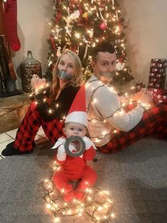 a man and woman are sitting in front of a christmas tree while holding a baby