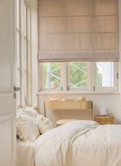 a white bed sitting under a window next to a desk with a lamp on it