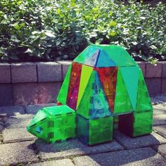 there is a colorful sculpture on the ground next to some plants and bushes in front of a brick wall