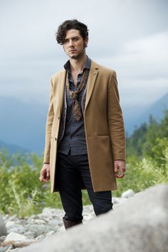 a man standing on top of a rocky hillside wearing a tan coat and black pants